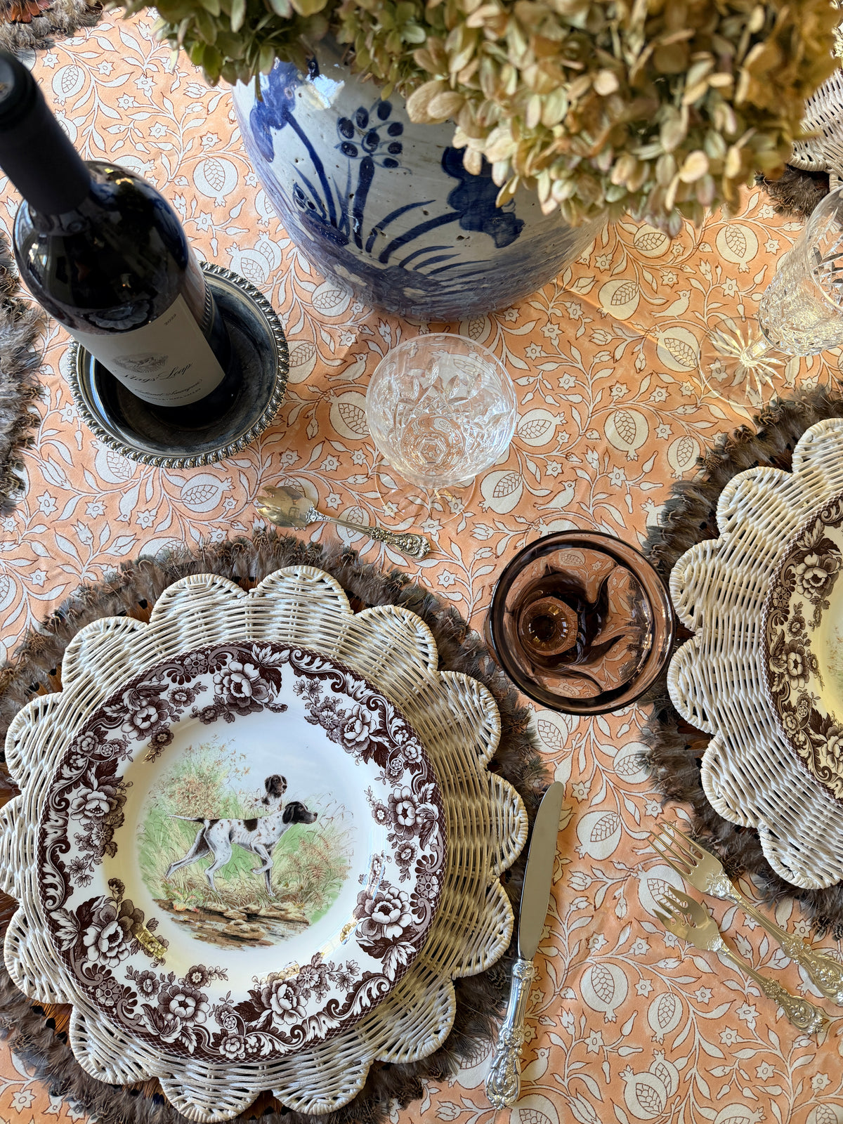 Pomegranate Clay hand block printed cotton tablecloth set with silver forks, a scalloped wicker charger and a brown transferware plate