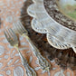 Close view of the Pomegranate Clay block print tablecloth pattern beside two ornate silver forks and a wicker charger