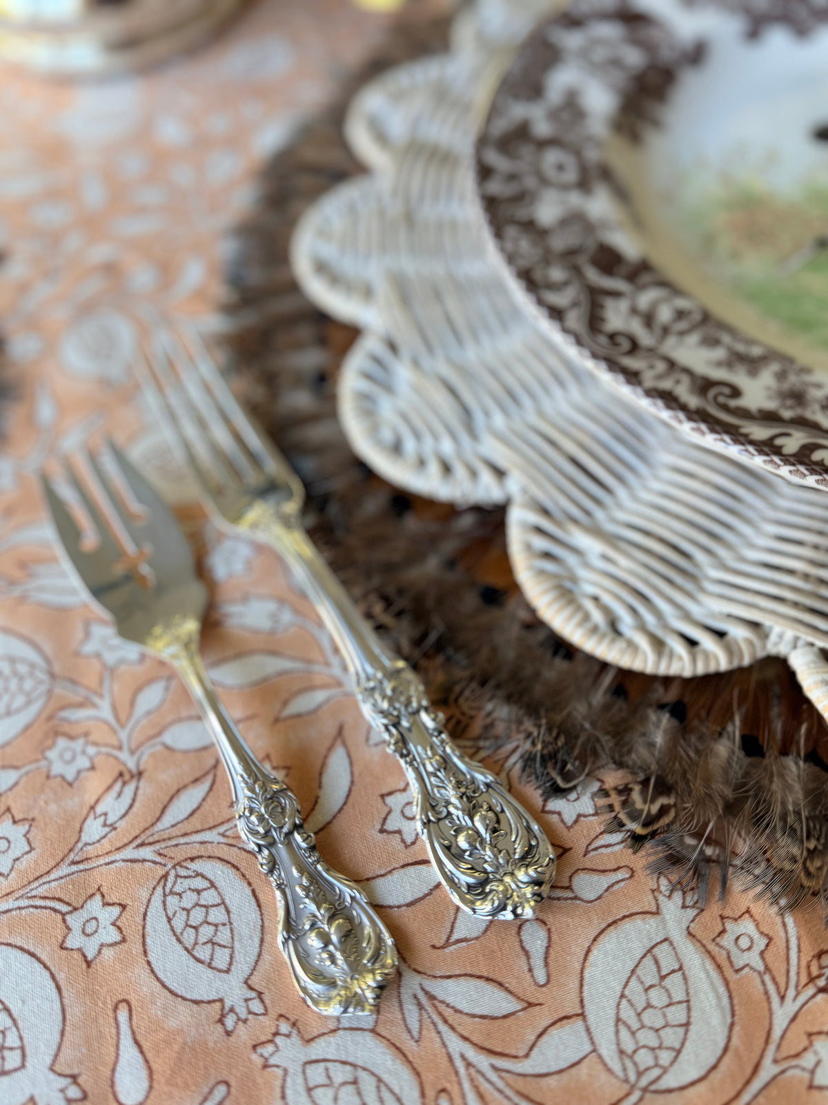 Close view of the Pomegranate Clay block print tablecloth pattern beside two ornate silver forks and a wicker charger